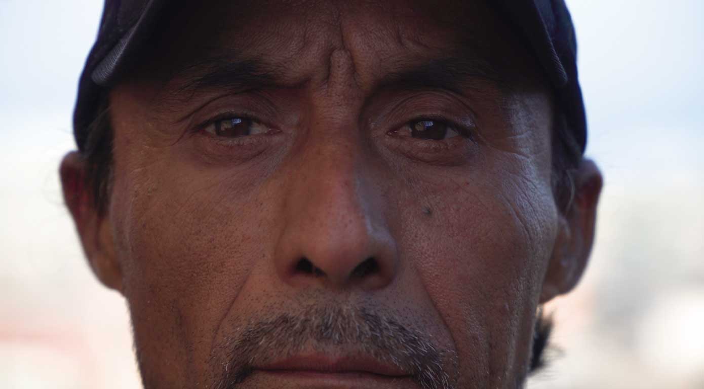 Rostro de una persona de tez morena, con un bigote delgado mirando de frente a la cámara, con un jockey y la ciudad de Antofagasta detrás