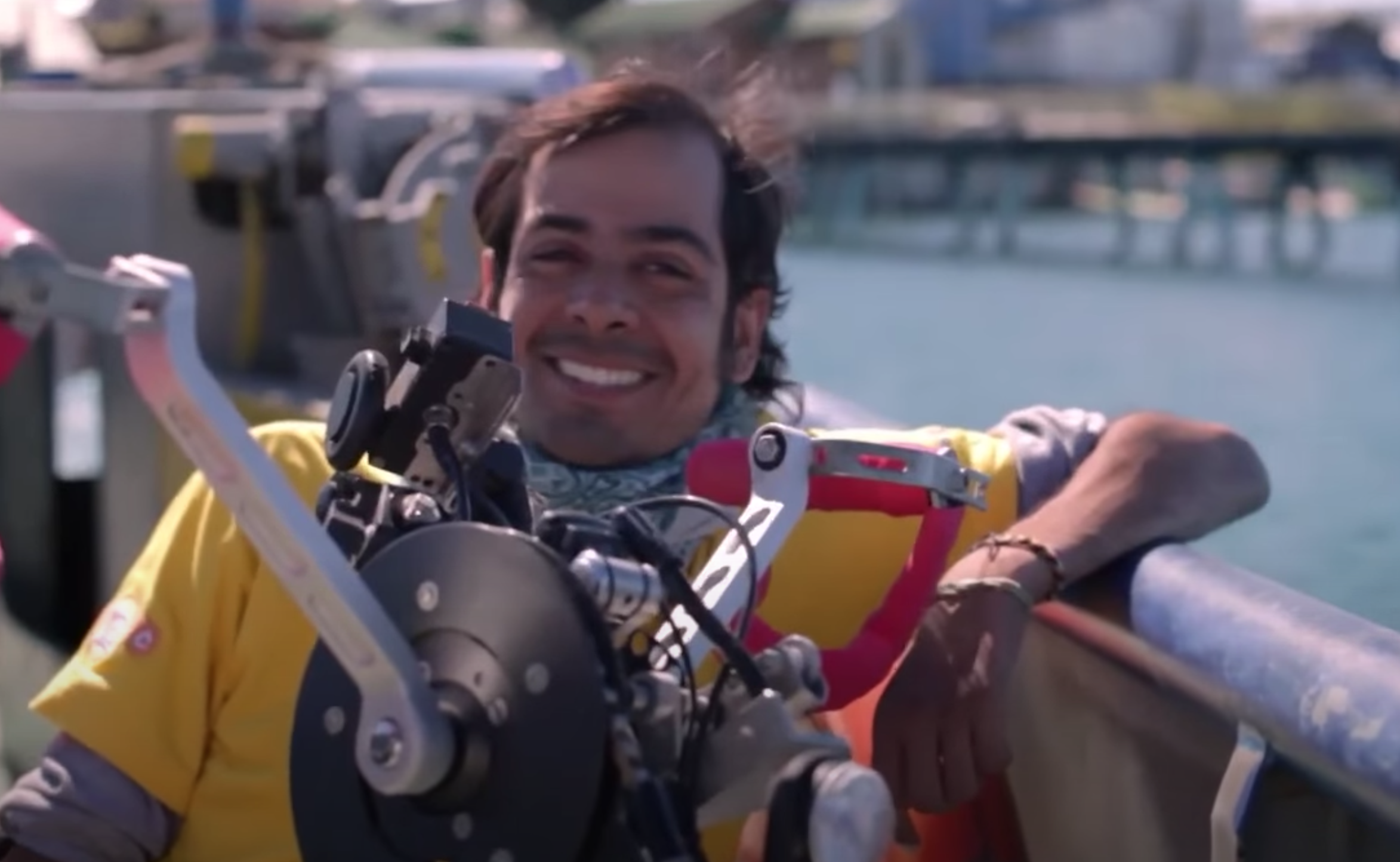 Felipe, persona con discapacidad, en silla de ruedas, está apoyado con su brazo izquierdo en una baranda. Atrás se ve un muelle y la orilla del mar