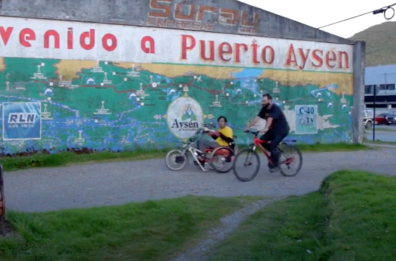 Dos personas, una en bicicleta y otra en silla de ruedas pasando frente a un mural que dice "bienvenido a Puerto Aysén"