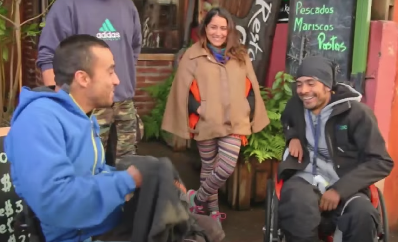 Dos personas en silla de ruedas sonriendo, una mujer sonriendo entre ellos y un joven parado al lado, afuera de un restaurant despidiéndose. El lugar es en el sur de Chile