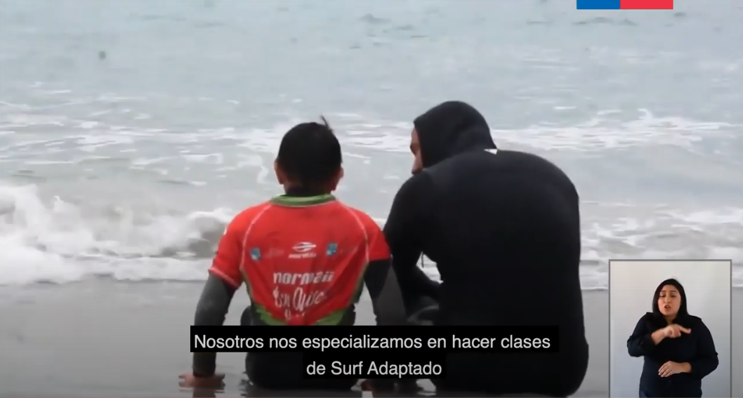 Foto con dos personas sentadas a la orilla del mar, un niño y un adulto, con trajes de surf conversando mirando el mar