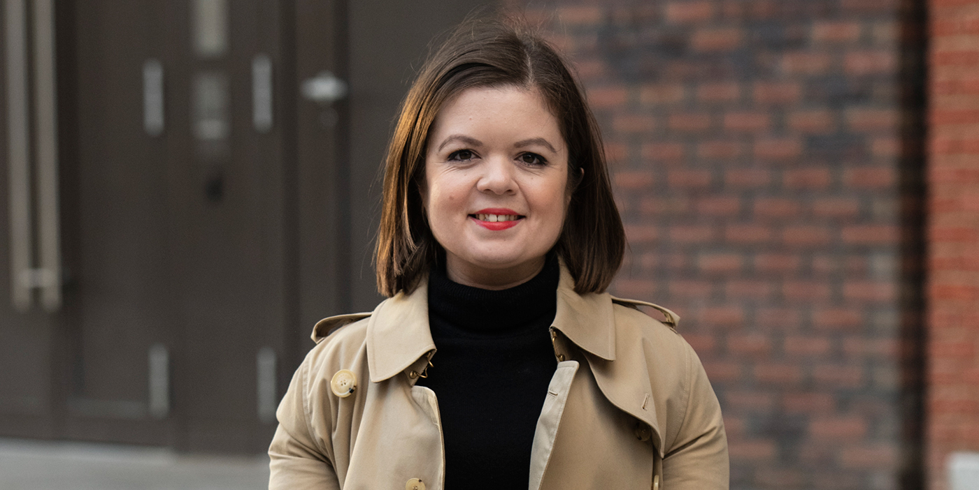 Foto de mujer de frente, sonriendo, con una chaqueta café y un chaleco negro