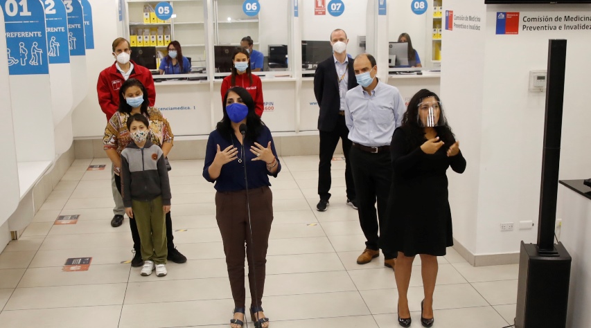 En la foto están 8 personas de pie, con sus mascarillas y distancia social, ordenadas en tres filas, al centro está la Ministra Rubilar hablando, a su izquierda tiene a la interprete de señas, detrás de ellos está una madre con su hijo y al lado el Ministro Monckeberg, al fondo están tres personas de pie. Esto en un pasillo de atención a público con tres ventanillas de atención.